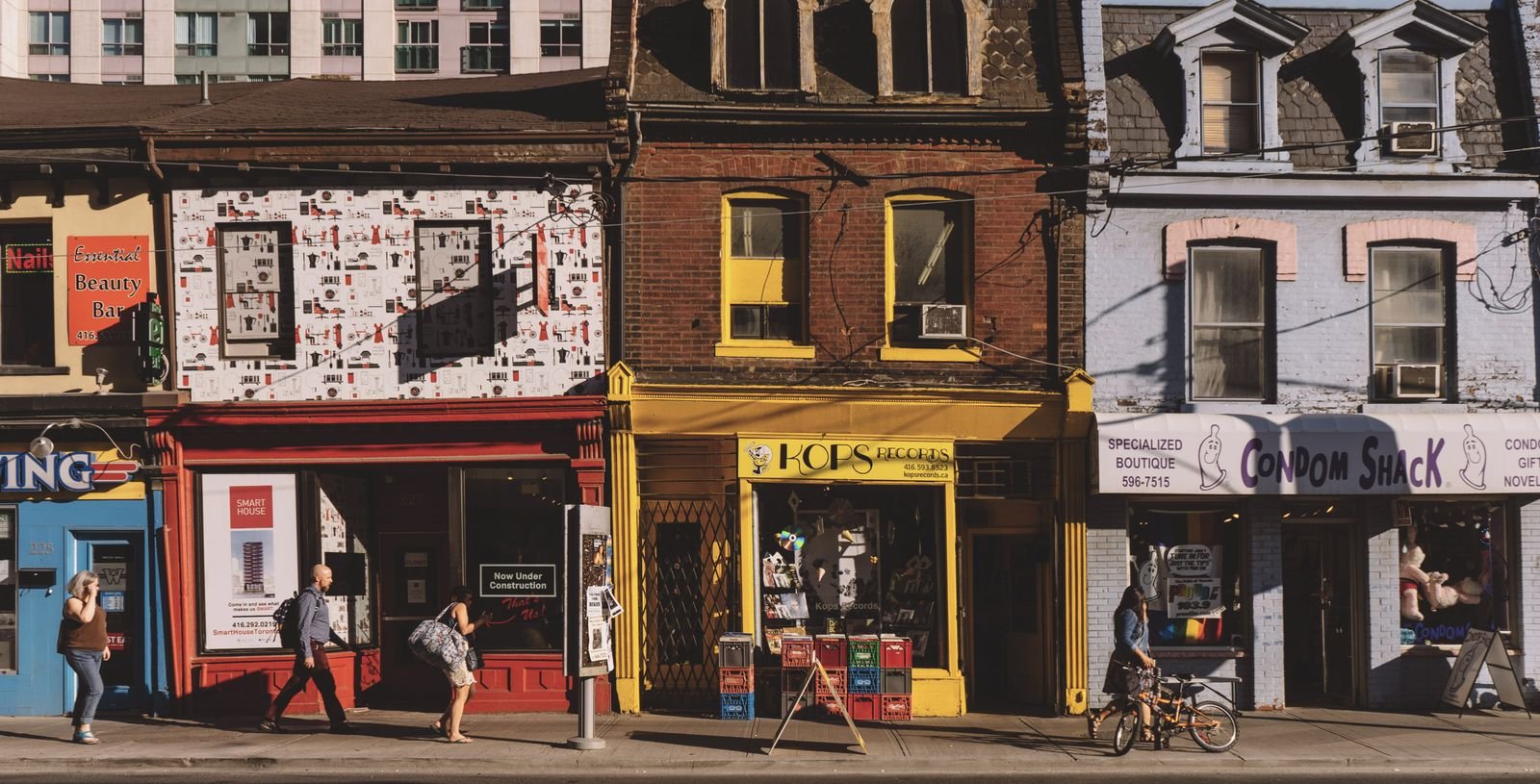 Pedestrians walking past a row of small storefronts on a sunlit street