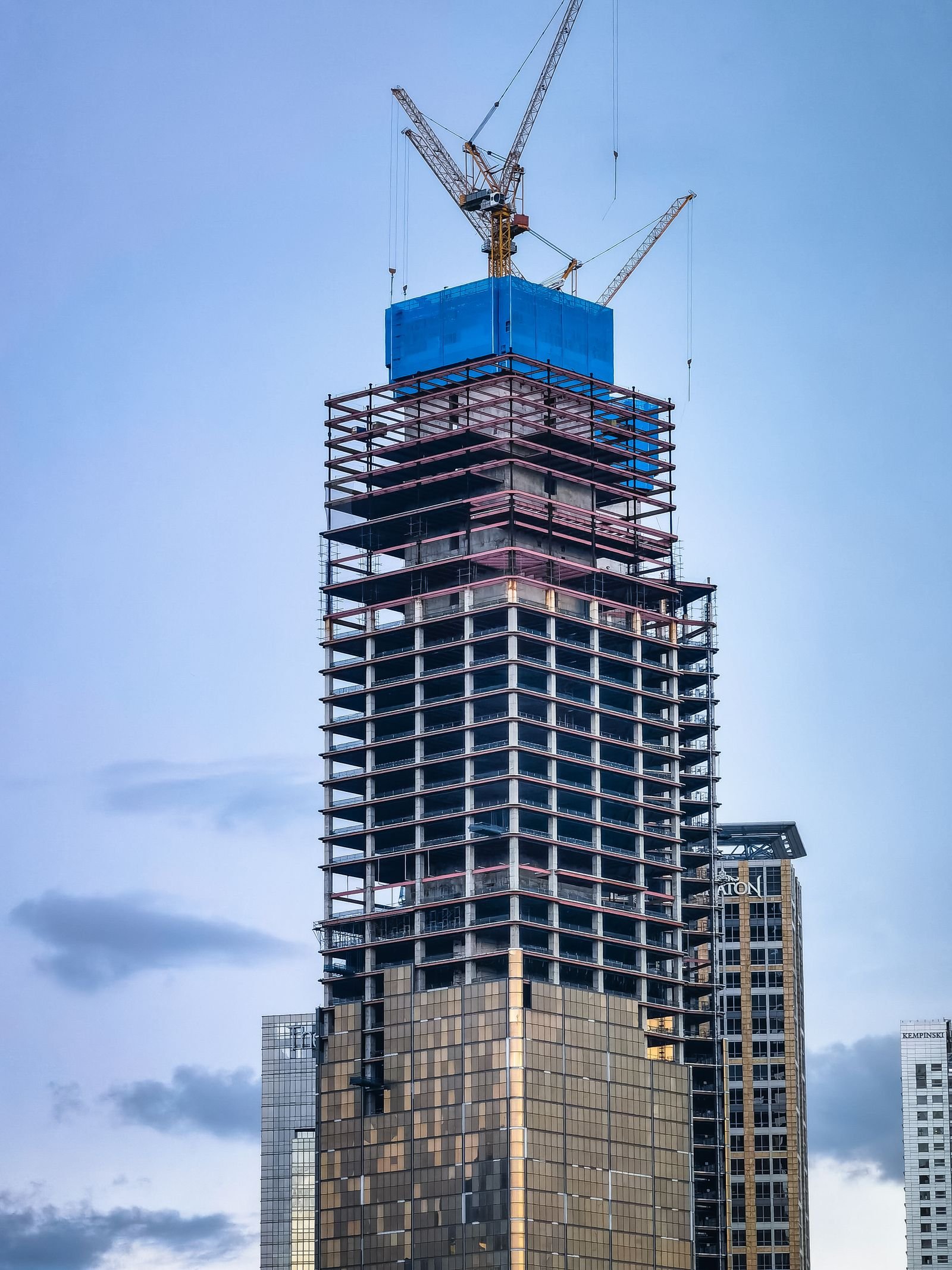 Tall glass skyscraper still under construction, framed by tower cranes